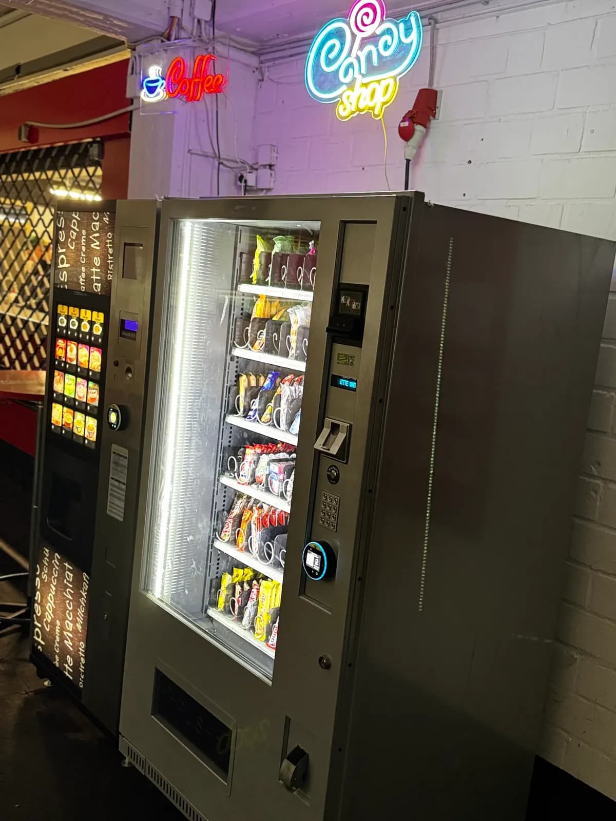 Kaffeeautomat und beleuchteter Snackautomat vor einer weiß gestrichenen Ziegelwand mit Leuchtschriften
